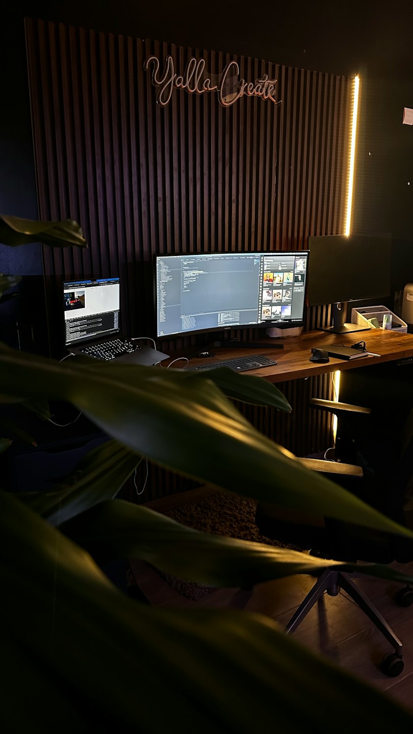 Modern desk with computers and green plant in a dark office.